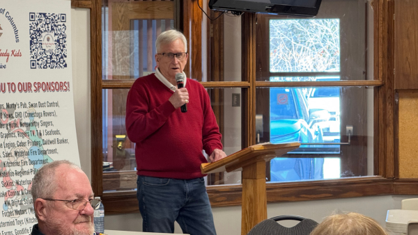 White-haired man in a red sweater and jeans, speaking into a microphone