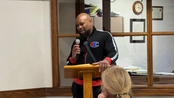 Black man in a workout jacket speaks into a microphone at a podium.